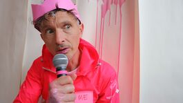 A white man with short grey hair is sat down and talking into a microphone. He’s wearing a pink paper crown on his head and a pink waterproof jacket with a sticky note on it that says ‘ME’. Behind him a pale pink liquid is dripping down the wall. The same liquid is also on his jacket, hand and lips as well.  
