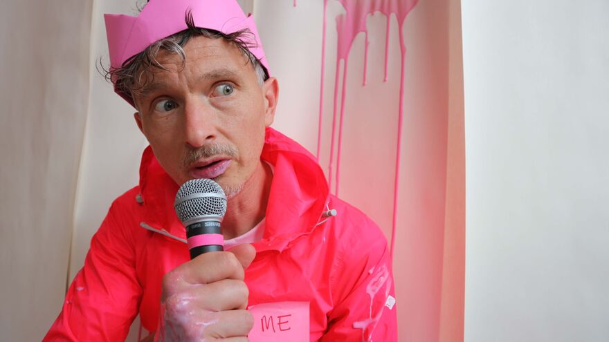 A white man with short grey hair is sat down and talking into a microphone. He’s wearing a pink paper crown on his head and a pink waterproof jacket with a sticky note on it that says ‘ME’. Behind him a pale pink liquid is dripping down the wall. The same liquid is also on his jacket, hand and lips as well.  