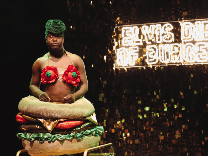 Francis, a black man, is wearing a 'drag burger' costume, including a bra with tomato shaped cups and a glittering burger dress. A neon sign behind reads 'Elvis Died of Burgers'