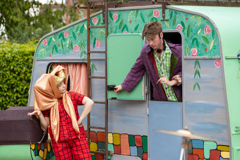 A man is opening his caravan door to greet the queen who is standing outside wearing a disguise.