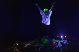 Jack, a white non-binary person with bright green hair and wearing a white t-shirt stands with their back to the camera under UV light, doing a morning stretch. They are surrounded by trinkets; a tiny pink flamingo, a book, leaves, a water bottle, a shiny pink love-heart shaped disco purse, and other nondescript things.
