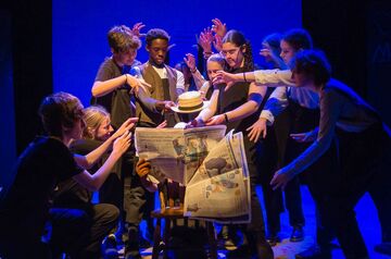 Group of young people gathered around man reading newspaper (made from props) reaching towards him, on a stage