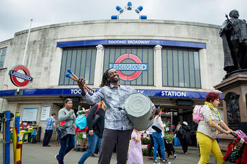 A man carrying a large drum outside of Tooting Underground Railway station.