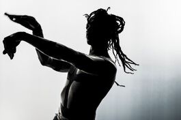 The Black and White Image Shows Bawren Tavaziva Standing With His Arms Outstretched And Making A Signature Gestural Curve With His Hands. Bawren Tavaziva Is A Man Of African Heritage, He Is Wearing His Hair Half Tied Up In Loose Braids And Has His Bare Torso And Arms Painted In Thick Dark Lines.