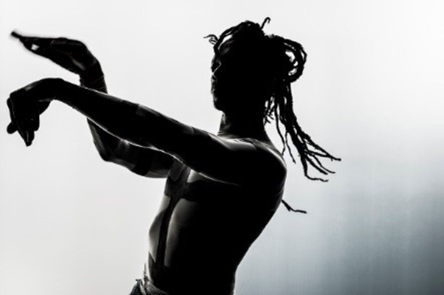The Black and White Image Shows Bawren Tavaziva Standing With His Arms Outstretched And Making A Signature Gestural Curve With His Hands. Bawren Tavaziva Is A Man Of African Heritage, He Is Wearing His Hair Half Tied Up In Loose Braids And Has His Bare Torso And Arms Painted In Thick Dark Lines.