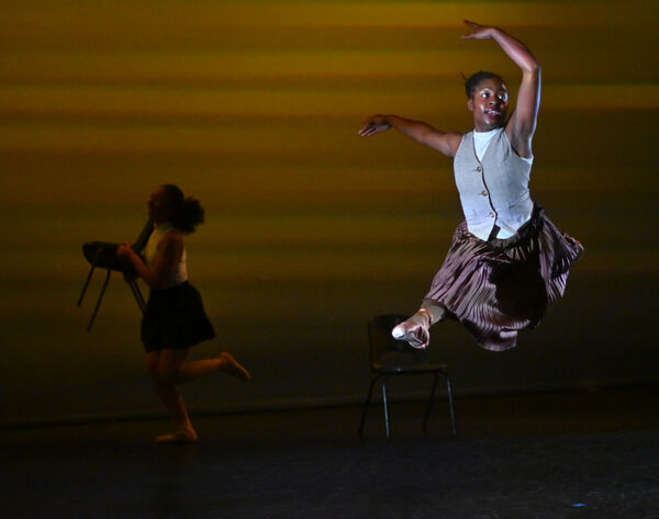 Black woman in grey vest and brown skirt jumping in the air gracefully on a dark stage. Another woman posing in the darkness behind her. 