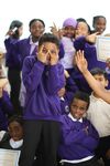 Boy with brown skin and hair in purple uniform, making face at camera with hands over face, more children smiling and posing in background