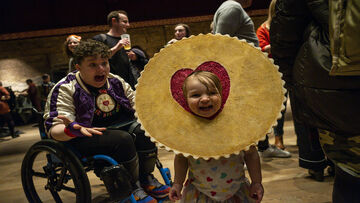 A young girl with a large (fake) jammy dodger around her face. Behind her a boy in a wheelchair can't believe his eyes. 