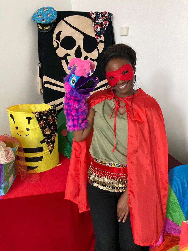Woman standing and wearing a red cape and mask, holding a purple and pink bird puppet. Background is a colourful prop box and large pirate themed cloth.