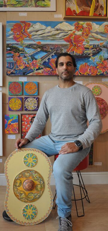 man sitting down in front of paintings on wall with a shield shaped sculpture resting against his leg