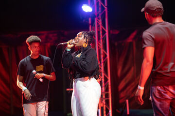 Black woman in black shirt and white trousers singing into microphone on stage, two men turned to face her on either sides of stage