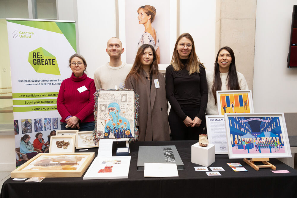 Five people stand together behind a table with their artwork on. Behind them is a pull-up banner with Re:Create on