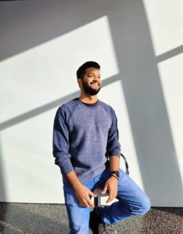 Man with brown skin and black hair, wearing blue jumper and jeans, sitting on a stool in a bright sunny room, smiling