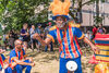Black main in colourful blue uniform and orange feathers in hat talks animatedly, outside under trees. Other people standing around behind him. 