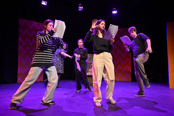 Five young people on stage, taking large steps, holding scripts