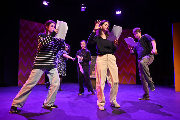 Five young people on stage, taking large steps, holding scripts