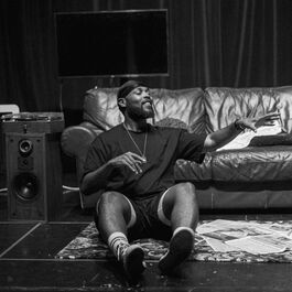 A black and white photo of a young black performer sitting on a patterned rug in front of a sofa, with his eyes closed and gesturing with his hands.