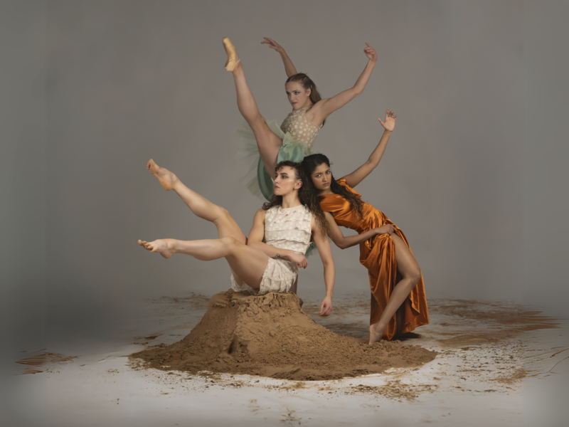 Three women, two are white and one has brown skin, surround a pile of sand against a grey background in a variety of dynamic balletic positions. 