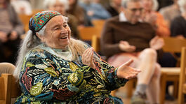 An elder woman wearing a leaf-patterned top clapping, in the background others are also clapping.