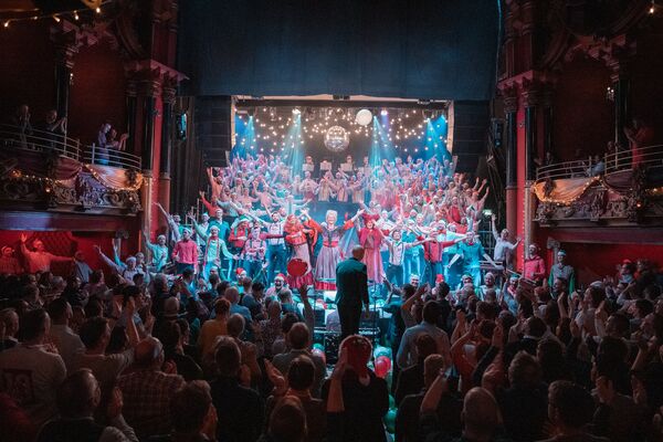 A finale moment featuring dozens of performers on stage with their arms raised as an audience applauds