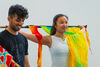 South East Asian woman in white vest top, holding colourful flag behind her shoulders. Standing next to South Aian man in black t-shirt, looking down and smiling 
