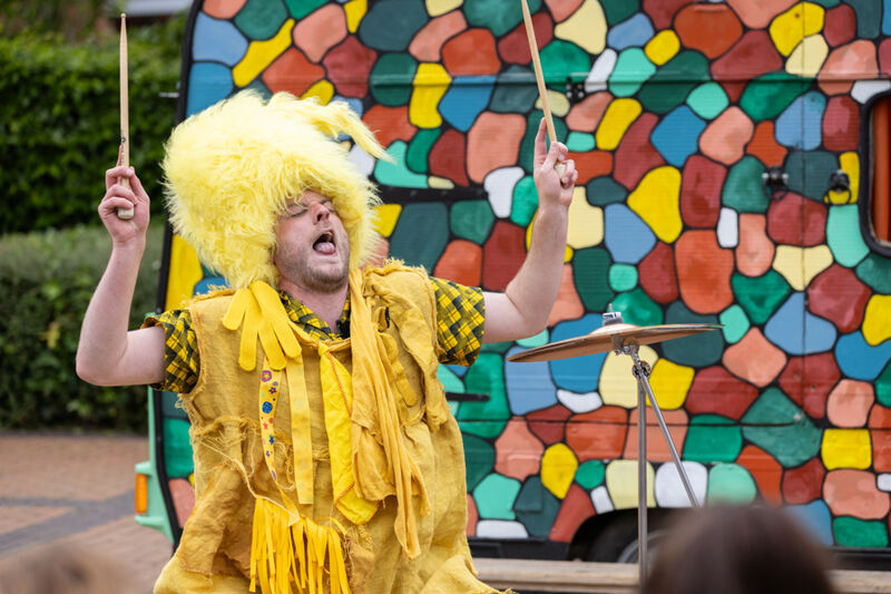 A troll dressed entirely in yellow is drumming and singing outside a brightly painted caravan.