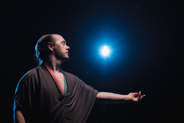 A male dancer stands in a dramatic pose, arm extended gracefully, with eyes closed. A bright blue spotlight shines behind them, creating a striking contrast against the dark background.
