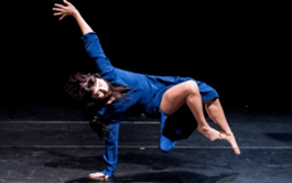 A woman with dark hair wearing a blue dress suspends herself from the floor of a dark stage with one arm, the other is raised above her and her legs are raised off the floor.