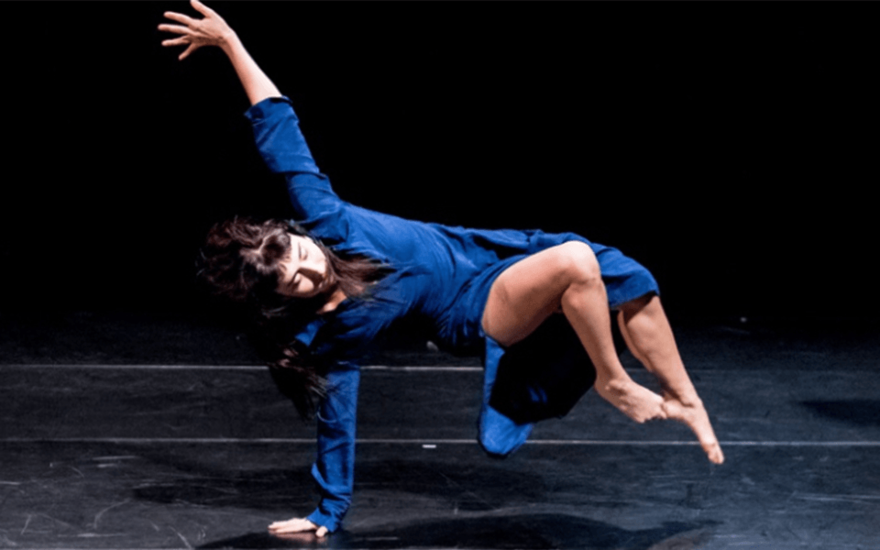 A woman with dark hair wearing a blue dress suspends herself from the floor of a dark stage with one arm, the other is raised above her and her legs are raised off the floor.