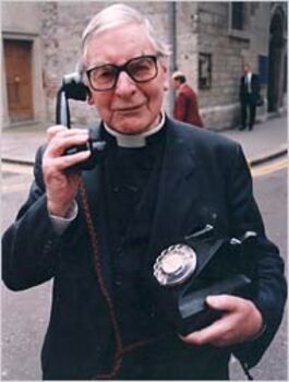 Image of Chad Varah holding a telephone and smiling. He is wearing his dog collar and black jacket.