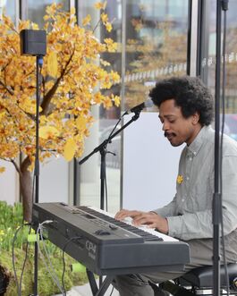 Black man in grey shirt playing keyboard, indoors, in front of tree with orange leaves