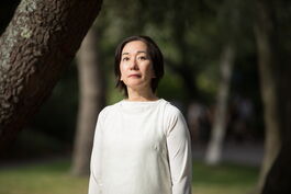 Chisato, a Japanese woman with a black bob. She stands in a white tunic dress in front of a blurred background of trees and greenery.