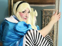 A grown up Woman in a blonde wig and a blue Alice in Wonderland dress with stripy tights looks cheekily to camera as she climbes through a looking glass.