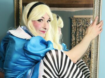 A grown up Woman in a blonde wig and a blue Alice in Wonderland dress with stripy tights looks cheekily to camera as she climbes through a looking glass.