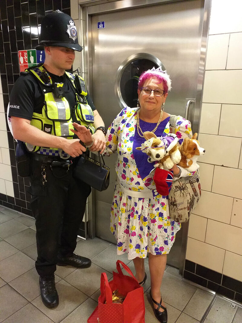 Performer dressed as Queen in tiara with toy corgis being handcuffed by police officer