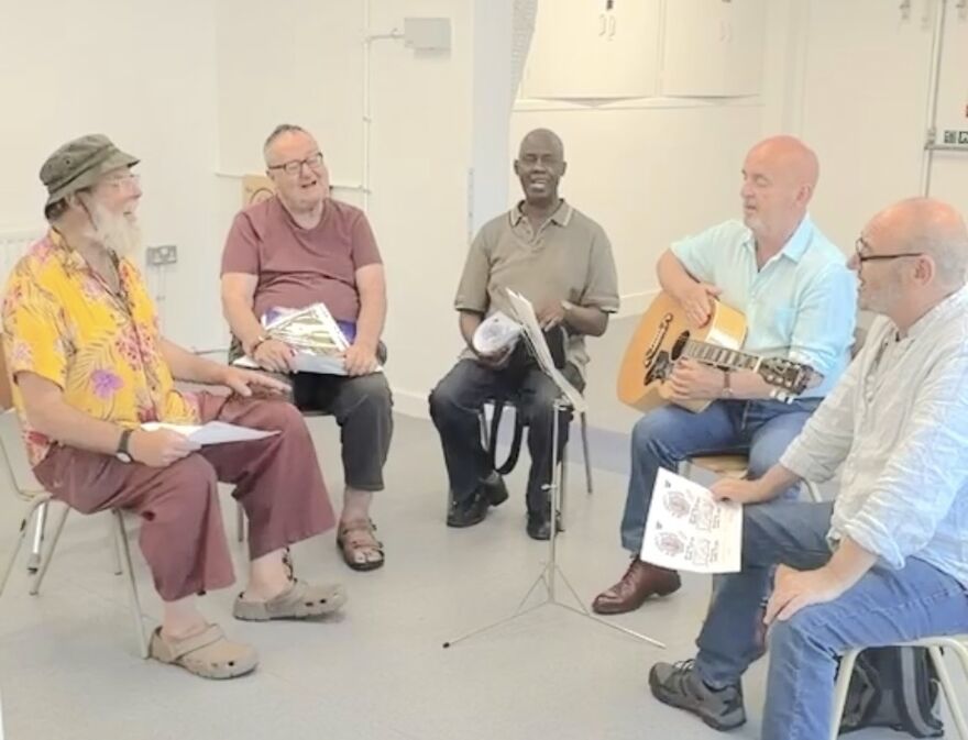 5 men sitting in a semi-circle, singing and looking happy