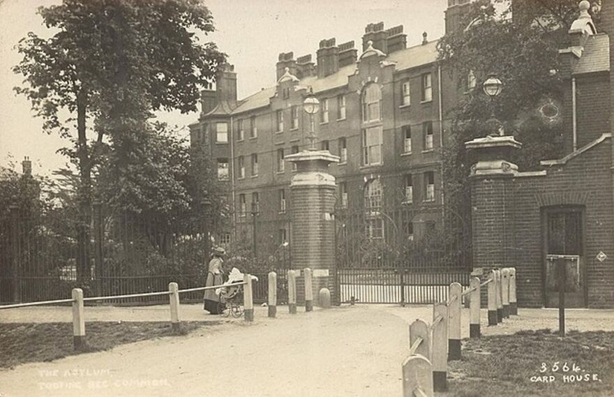 Image shows the outside of Tooting Bec Asylum with a woman pushing a pram walking past.