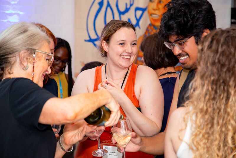 A person in an orange sleeveless top is smiling towards the right of the image at someone unseen, in front of them out of focus is another person with dark hair smiling at the person opposite them who is pouring a glass of champagne.