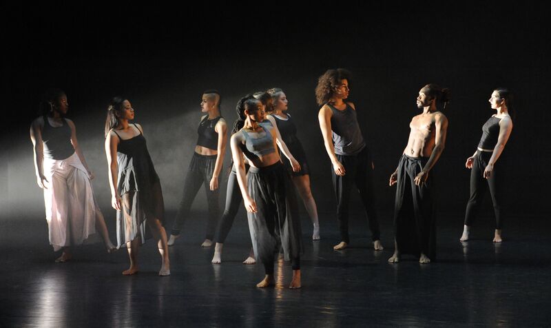 A group of dancers stand with their heads turned to the side and body tilted slightly. 