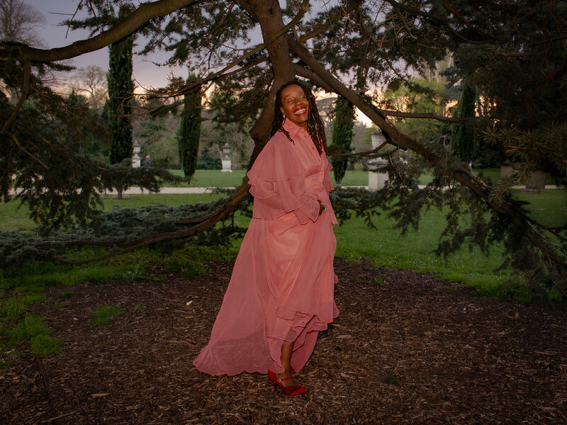 Publicity image of Singer, Melissa James wearing flowing pink vintage dress among trees, red shoes