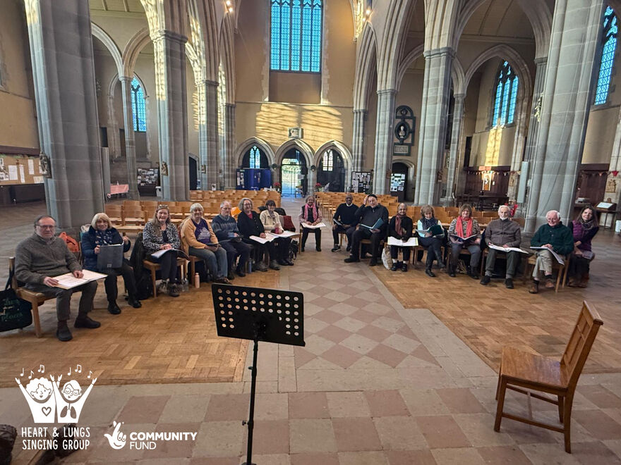 The friendly-looking and diverse members of Heart & Lungs Singing Group, seated, looking at the camera and ready to begin their singing session in their beautiful venue of All Saints Church, Tooting. 