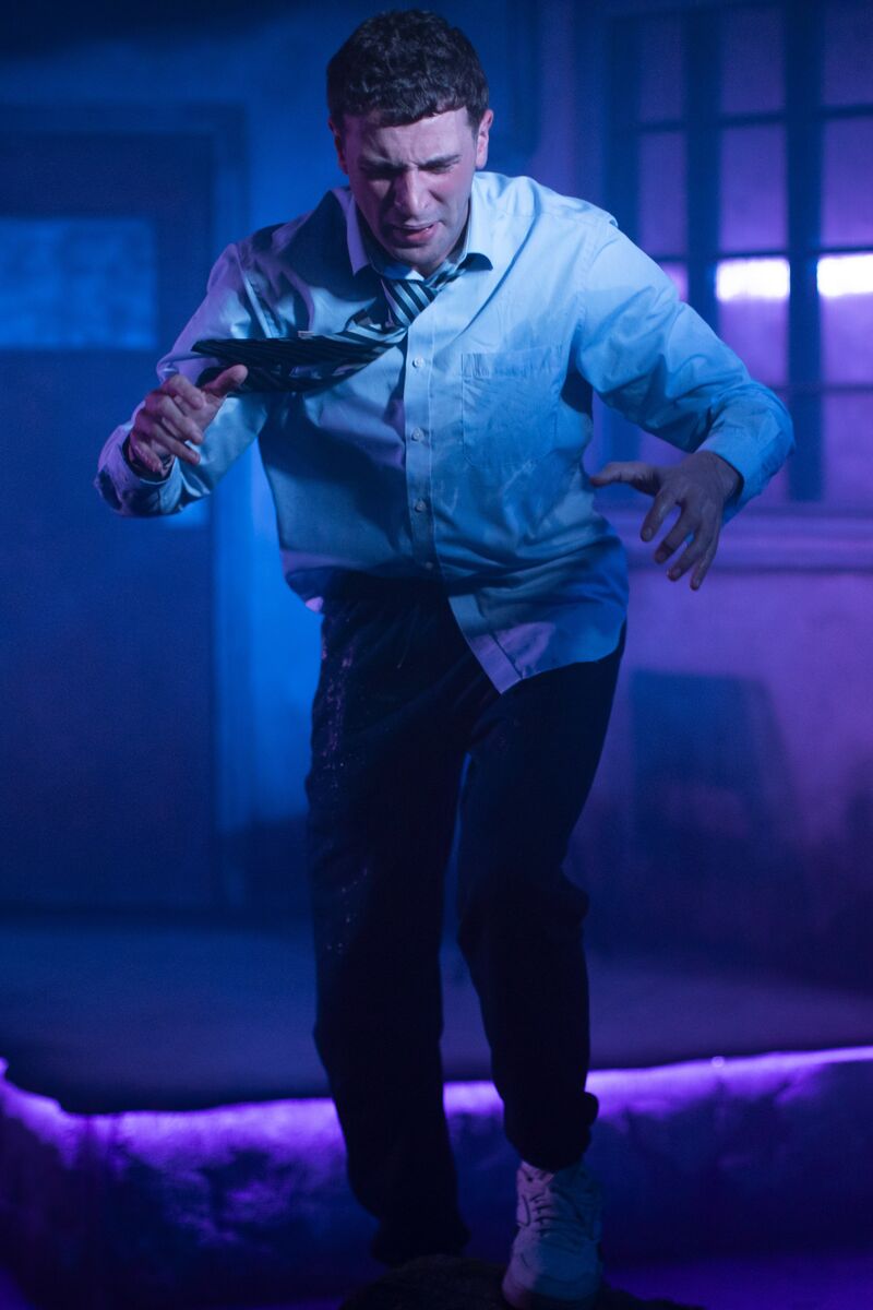 A man in a white shirt and dark trousers is photographed mid jump. He is grimacing. The lighting is purple and blue .