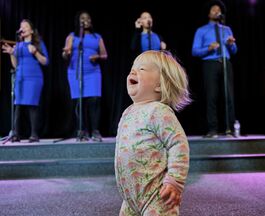 laughing toddler in foreground, 4 gospel singers dressed in royal blue in background behind microphones on stands.