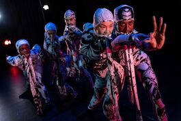 This image shows the five GREED dancers in their white and black body-fitting costumes with matching straps in their mouths. Dancer, Yanki is at the front of the image reaching her left arm out to hold dancer, Oraine's arm as he creates an OK sign with his left hand. All the dancers are looking towards Oraine's hand.