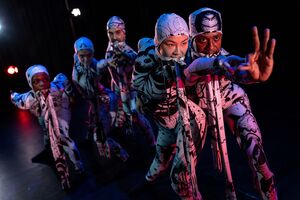 This image shows the five GREED dancers in their white and black body-fitting costumes with matching straps in their mouths. Dancer, Yanki is at the front of the image reaching her left arm out to hold dancer, Oraine's arm as he creates an OK sign with his left hand. All the dancers are looking towards Oraine's hand.