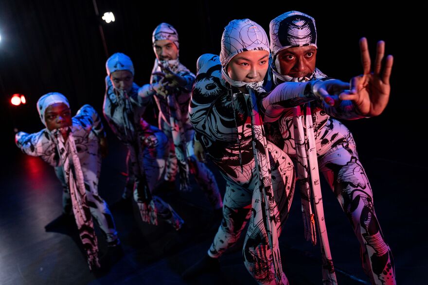 This image shows the five GREED dancers in their white and black body-fitting costumes with matching straps in their mouths. Dancer, Yanki is at the front of the image reaching her left arm out to hold dancer, Oraine's arm as he creates an OK sign with his left hand. All the dancers are looking towards Oraine's hand.