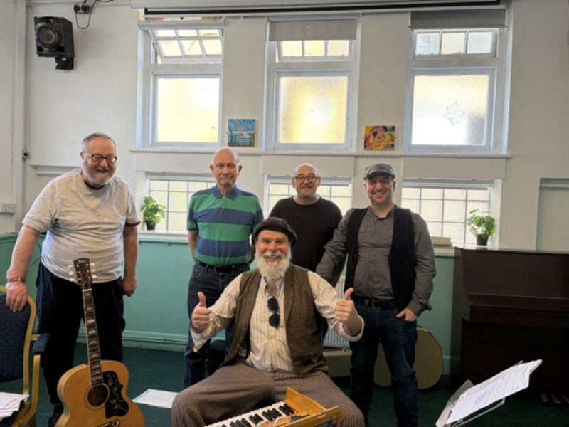 four men standing with one in front that is sitting at a wooden indian harmonium, thumbs up. all looking-at and smiling-at the camera