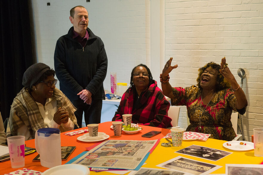 Three women and one man around a table and one of the women is singing