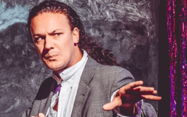 A white man with long curly hair tied back in a ponytail is wearing a suit and extending out his arms as if ready to cast a spell. He looks concerned, as if he’s trying to ward something off. Behind him is a window painted black and a metallic red and purple curtain. The whole image has a paint-like quality to it, as if it’s been put through a retro filter on photo-booth to make it appear more like a poster.