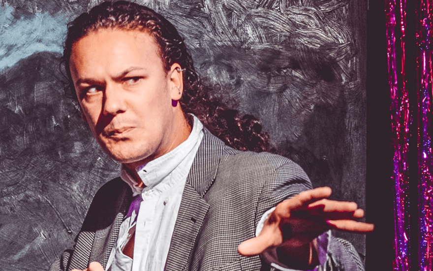 A white man with long curly hair tied back in a ponytail is wearing a suit and extending out his arms as if ready to cast a spell. He looks concerned, as if he’s trying to ward something off. Behind him is a window painted black and a metallic red and purple curtain. The whole image has a paint-like quality to it, as if it’s been put through a retro filter on photo-booth to make it appear more like a poster.
